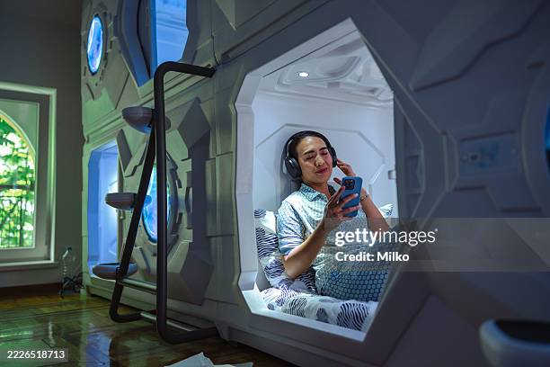 mid adult woman using smartphone while relaxing in capsule hostel - capsule hotel stock pictures, royalty-free photos & images
