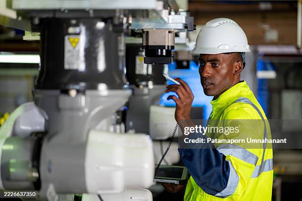 un ingeniero negro profesional con ropa de trabajo protectora usa una tableta digital para evaluar la calidad de un brazo robótico dentro de una fábrica de alta tecnología. centrada en la precisión y el cumplimiento, la inspección garantiza que el pro - ingeniería mecánica fotografías e imágenes de stock