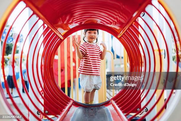 joyful little asian girl having fun playing at jungle gym in an outdoor playground on a sunny day. smiling and looking at camera. healthy family lifestyle. happy childhood. fun and healthy physical outdoor activities for kid - spielgerät stock-fotos und bilder