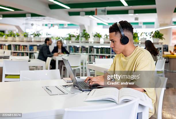university student at the library using headphones while studying on his laptop - dissertation stock pictures, royalty-free photos & images