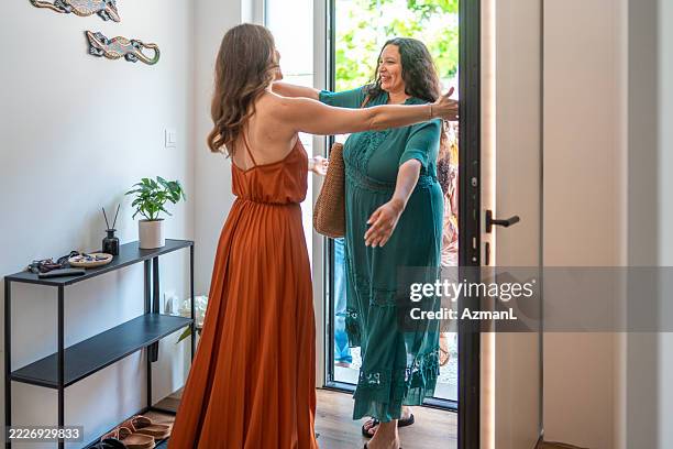 smiling mid adult women greeting each other in bright home entrance wearing elegant dresses - party host stock pictures, royalty-free photos & images