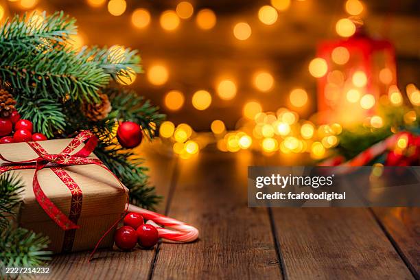 cadeau de noël enveloppé de papier kraft et de ruban rouge sous le sapin de noël dans une atmosphère festive chaleureuse avec des lumières de noël défocalisées en arrière-plan - sapin de noël photos et images de collection