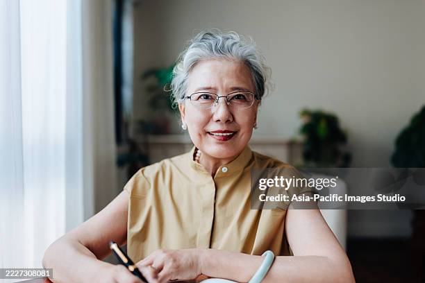 portrait of asian senior woman sitting on a sofa, looking at camera, while studying on laptop in the living at home. e-learning concept. senior people and retirement concept. - chinese ethnicity stock pictures, royalty-free photos & images