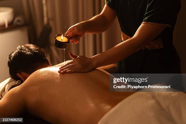il terapeuta tailandese versa l'olio caldo di una candela da massaggio sulla schiena di un uomo durante una rilassante sessione termale. il tocco delicato, la luce dorata e l'atmosfera curativa evocano un profondo relax. - aromaterapia foto e immagini stock