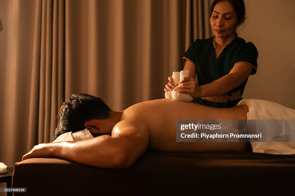 Thai therapist performing herbal compress massage on a man back in a calming spa environment. Traditional technique promotes deep muscle relief, stress reduction, and holistic wellness.