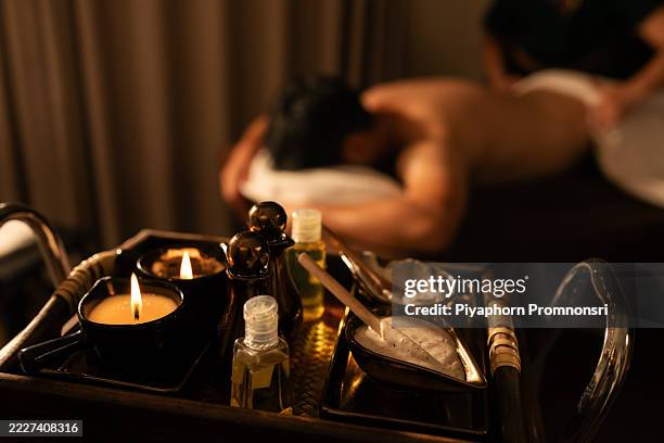 traditional thai massage setup with herbal oils, aromatic candles, and body cream in a luxury spa. male client receiving back massage in blurred background, creating a calm and relaxing ambiance. - hand massage stock pictures, royalty-free photos & images