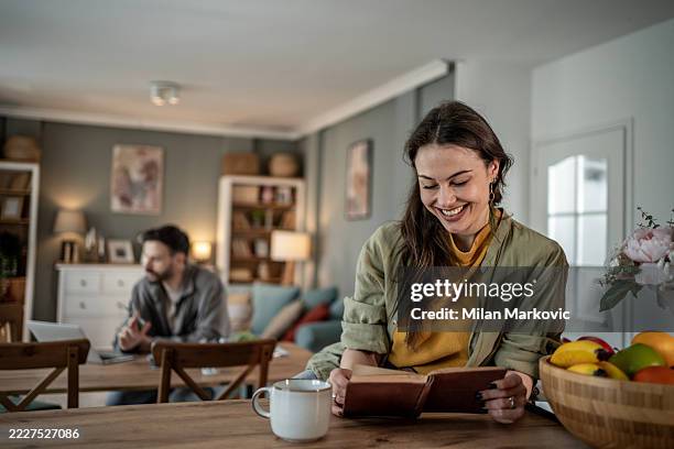 young woman reading a book and smiling at home while man works on laptop - self improvement stock pictures, royalty-free photos & images