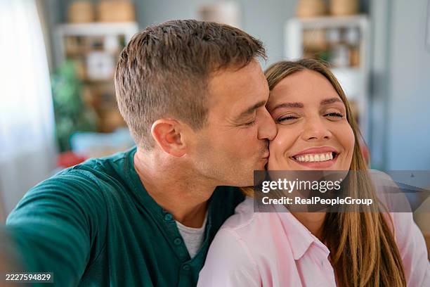 glückliches paar, das ein selfie macht, während es sich zu hause küsst - wange stock-fotos und bilder