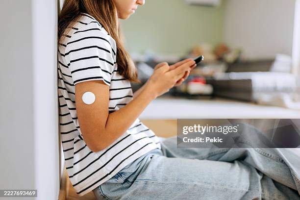 teenage girl with diabetic sensor attached on her arm using smart phone while sitting on floor at home - type 1 diabetes stock pictures, royalty-free photos & images