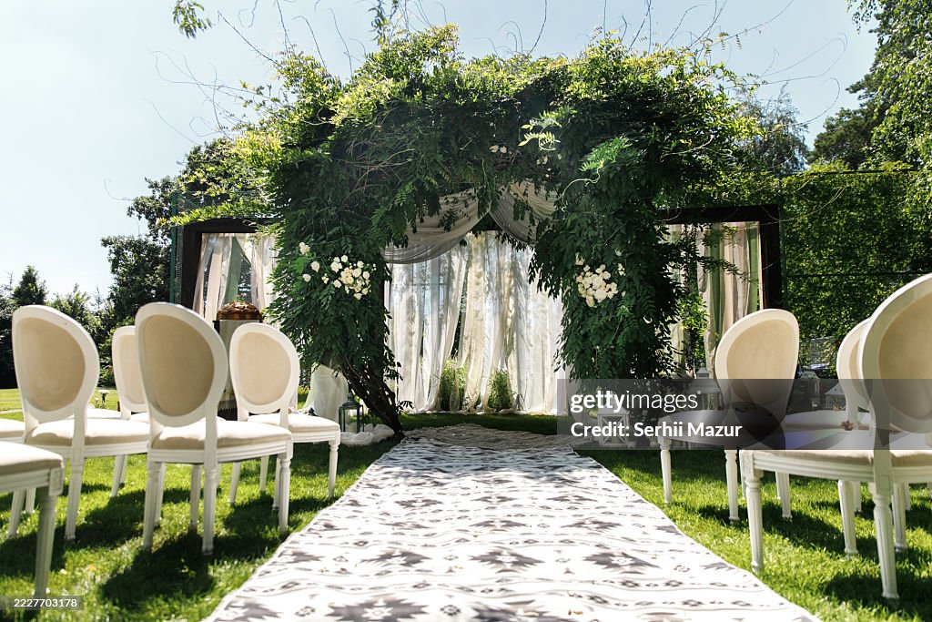 Elegant outdoor wedding setup featuring a floral arch and white chairs in a lush garden during the daytime.