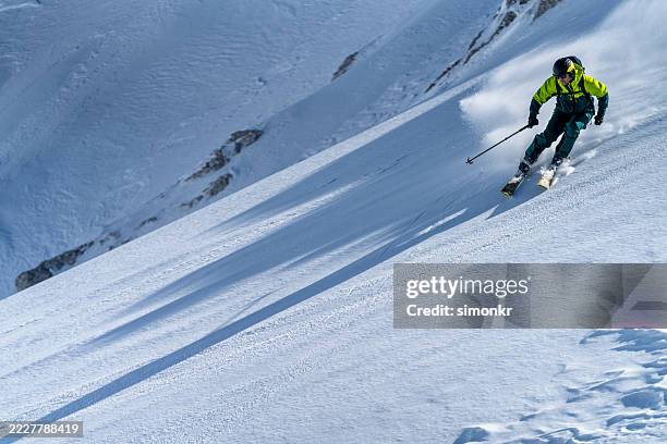 athletic skier in bright green jacket descending pristine snow-covered mountain slope under clear sky - professional skier stock pictures, royalty-free photos & images