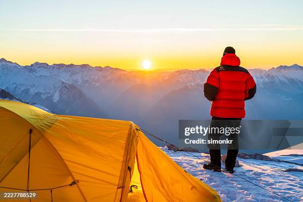 bergabenteurer in roter winterjacke, der den sonnenaufgang über verschneiten alpengipfeln in der nähe des gelben zeltes beobachtet - basislager stock-fotos und bilder