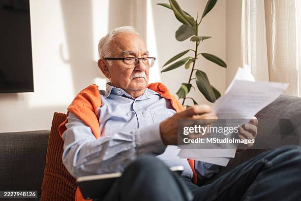senior man reading documents while sitting on a sofa in bright sunlight - wise old man stock pictures, royalty-free photos & images