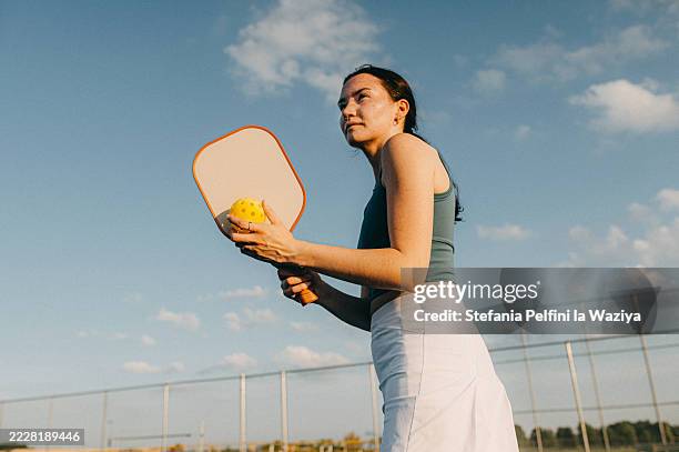a young woman stands on a tennis court, holding a paddle and a pickleball, ready to play under a clear sky. - racquet sport stock pictures, royalty-free photos & images