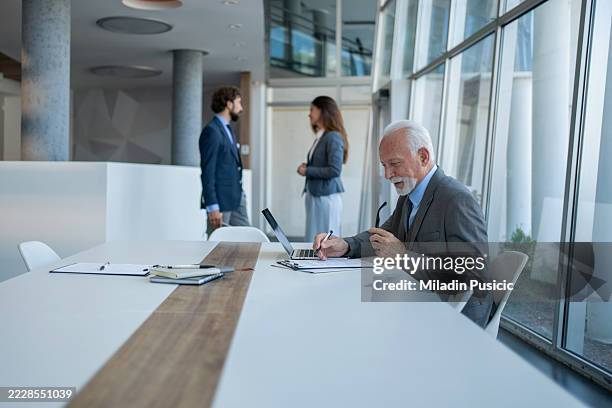 senior businessman working in modern office while colleagues are talking in background - hierarquia de corporação imagens e fotografias de stock