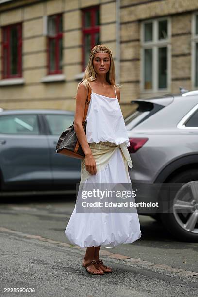 Guest wears a loose-fitting white sleeveless top with thin straps, paired with a voluminous white skirt featuring a gathered hem. A beige fabric is...