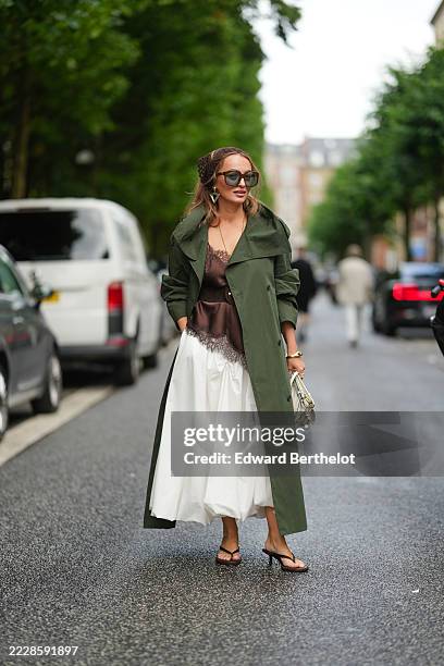 Guest wears oversized sunglasses with dark lenses and a headband. Earrings are visible. The outfit includes a dark green trench coat with a wide...