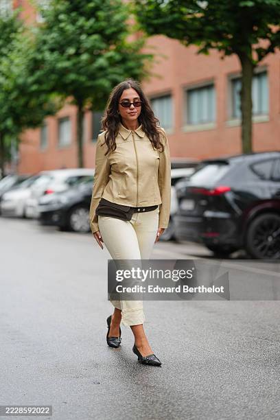 Guest wears a beige jacket with a pointed collar and front zipper closure, paired with cream-colored cropped pants. Dark sunglasses are worn, and a...
