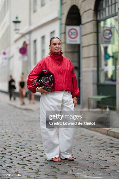 Guest wears a high-neck red jacket with black buttons and long sleeves. The jacket is paired with wide-leg white trousers. Accessories include gold...