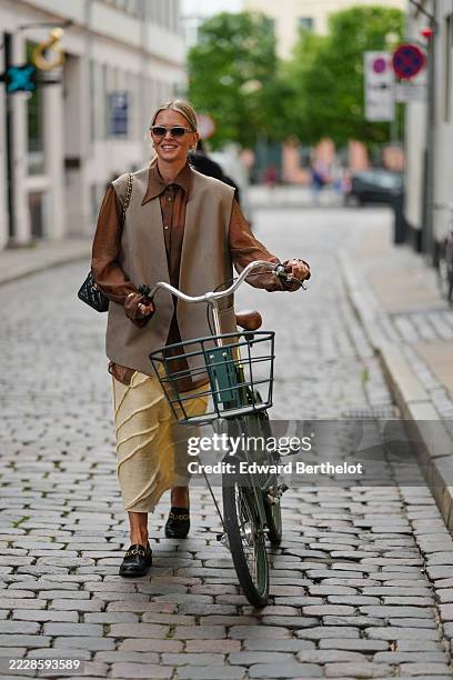 Guest wears sunglasses with a thin frame and light lenses. Hair is styled in a sleek, low bun. A sleeveless, oversized vest in a neutral tone is...