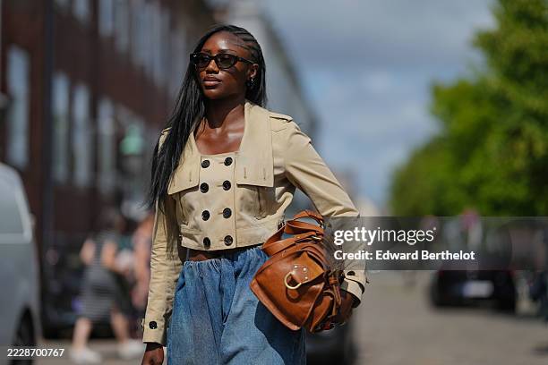 Guest wears a cropped beige trench coat featuring black buttons and epaulets on the shoulders. Blue high-waisted denim pants are worn, showcasing a...