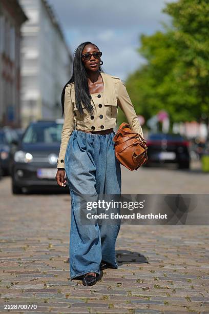 Guest wears a cropped beige trench coat with black buttons and epaulets, paired with high-waisted, wide-leg blue denim jeans. The outfit is...