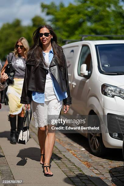 Guest wears sunglasses with dark lenses and frames. Hair is styled long and straight. A black leather jacket with a wide collar and button details is...
