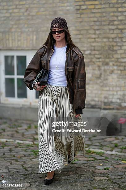 Guest wears a brown crochet beanie and rectangular sunglasses. A brown leather bomber jacket with visible stitching and a white crew-neck T-shirt...