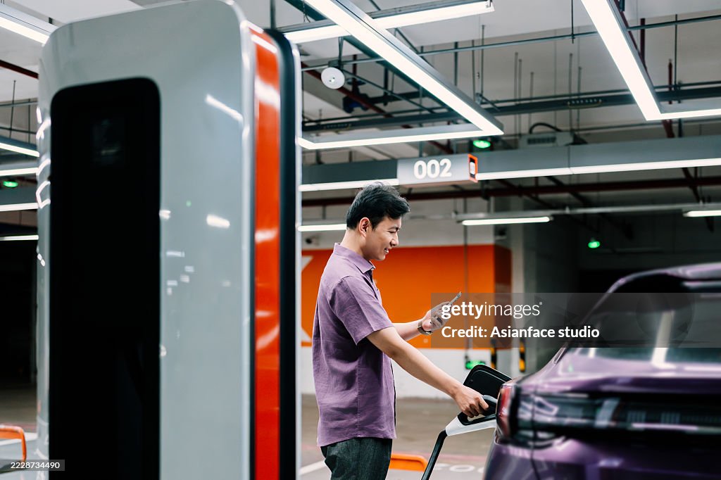 Orange and purple series. Side view of young Asian man plugging cable in electric car at a public underground charging station. Electric vehicle with charger at a public charging station.