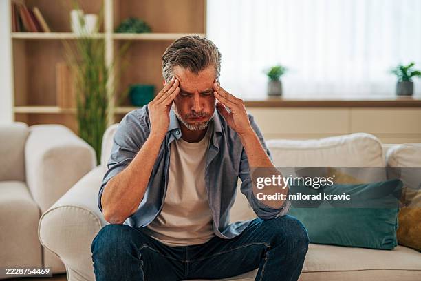 homme mûr souffrant de maux de tête alors qu’il est assis sur un canapé à la maison - mal de tête photos et images de collection