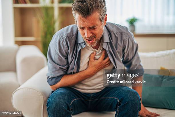 mature man suffering from chest pain while sitting on sofa - hartinfarct stockfoto's en -beelden
