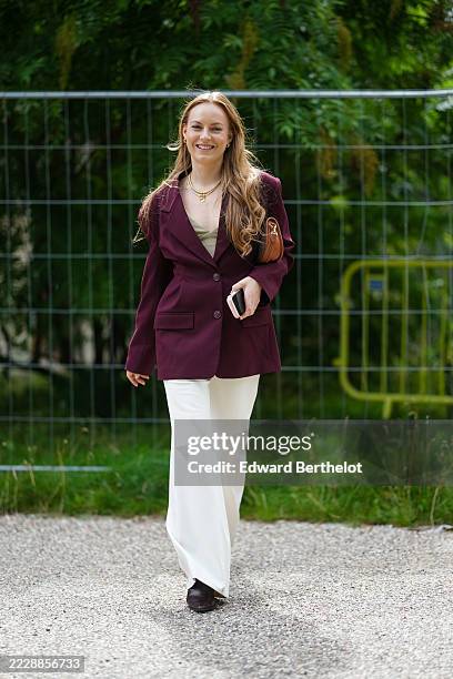 Guest wears long, wavy hair and a burgundy blazer with structured shoulders and two-button fastening. Underneath, a light-colored top is visible. The...