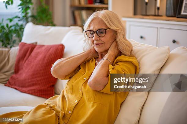 senior woman suffering from neck pain while relaxing on sofa - osteoporose imagens e fotografias de stock