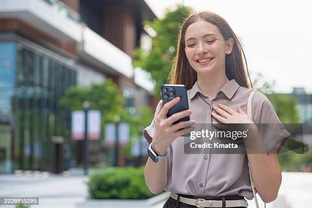 smiling young businesswoman holding a smartphone with happy expression feeling relieved. - hands on chest stock pictures, royalty-free photos & images