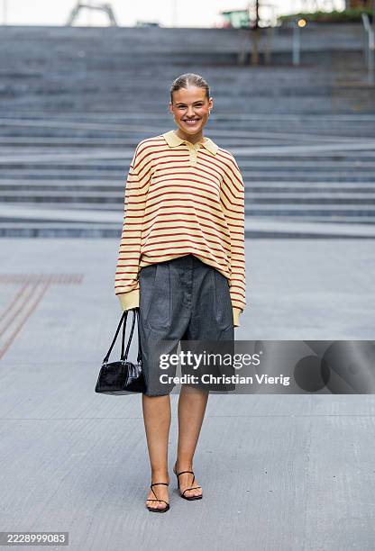 Guest wears beige striped oversized polo shirt with long sleeves, grey shorts, black bag outside Deadwood during Copenhagen Fashion Week day four on...