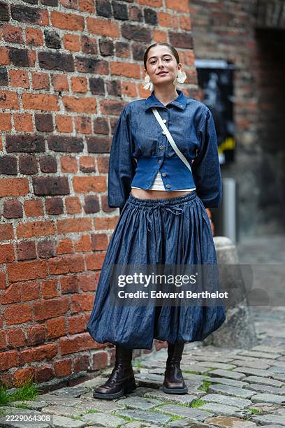 Guest wears dark brown hair slicked back into a center-parted low bun, large off-white floral drop earrings, and a white crossbody strap worn...