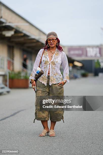 Guest wears a beige crochet skullcap embellished with small crystals, shoulder-length wavy dark red hair, rectangular brown acetate glasses with...