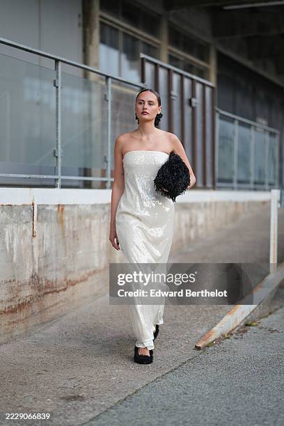 Guest wears a strapless, floor-length gown covered in white sequins, creating a shimmering effect. The hair is styled in a sleek, low bun. A black,...