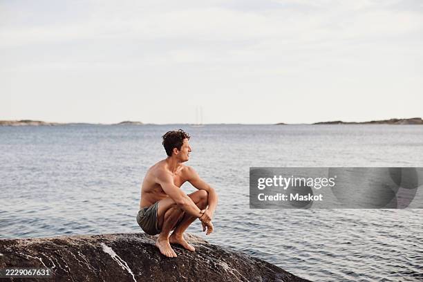 shirtless man looking away while crouching on rock near sea - lättklädd bildbanksfoton och bilder