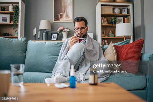 sick man sitting on sofa checking his temperature with thermometer - doente imagens e fotografias de stock