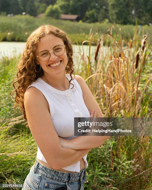 selbstbewusst lächelnde junge erwachsene frau mit brille, die in die kamera schaut, mit verschränkten armen im sommer draußen in der natur. - ärmellos stock-fotos und bilder