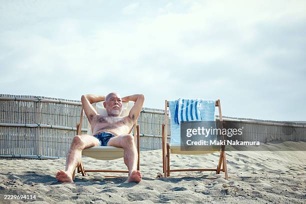 man stands alone in a deck chair on a quiet beach, staring into the distance at the sparkling sea. - liegestuhl stock-fotos und bilder
