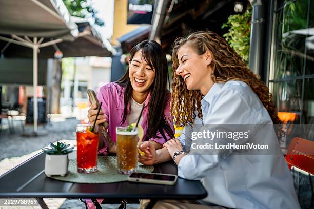 two young women laughing while looking at smartphone in outdoor cafe - terras stockfoto's en -beelden