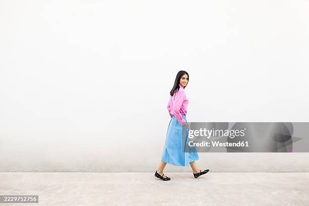 confident woman in pink shirt and blue skirt walking by a white wall - vestido-de-colores fotografías e imágenes de stock
