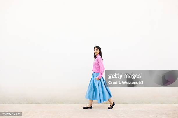 confident woman in pink shirt and blue skirt walking by a white wall - white skirt stock pictures, royalty-free photos & images