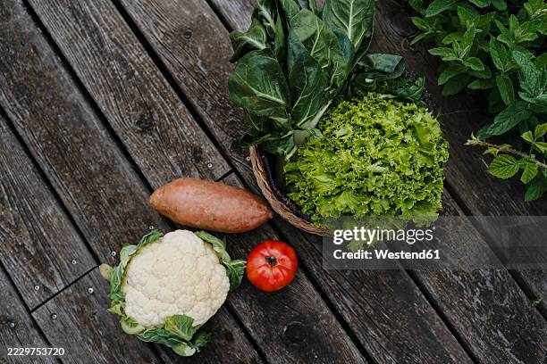 fresh vegetables including cauliflower, lettuce, sweet potato, and tomato on a wooden surface outdoors - crucifers stock pictures, royalty-free photos & images