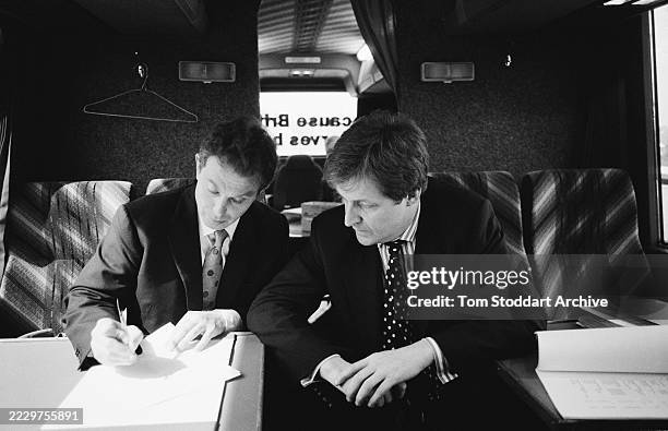 British politician Tony Blair, wearing a dark suit over a white shirt and a polka dot tie, writing beside British strategist Alastair Campbell,...