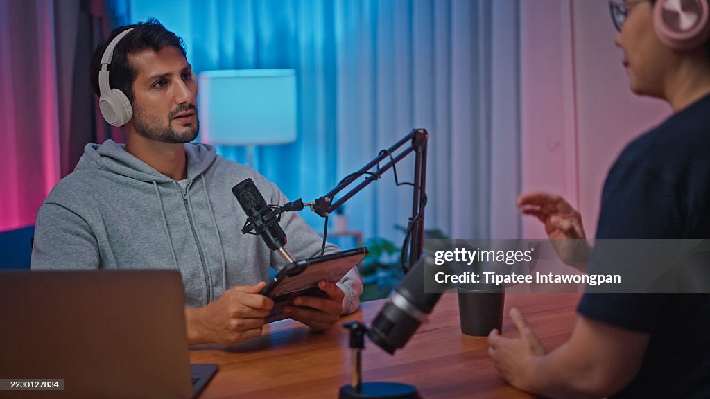 Young male and female podcasters wear headphones speaking in microphone and interview sit at desk with laptop at studio. Streaming live podcast concept.