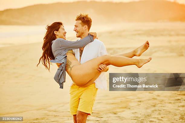 wide shot happy man carrying girlfriend on empty beach at sunset - fully unbuttoned stock pictures, royalty-free photos & images