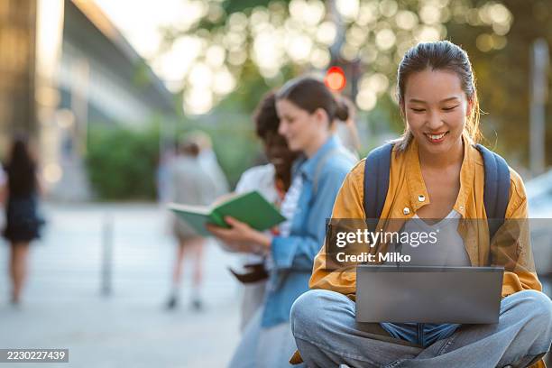 young woman using a laptop outdoors with friends studying in the background - universiteitsstudent stockfoto's en -beelden